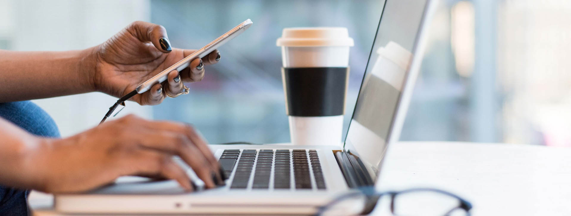 A woman holds a smartphone in one hand while typing on her laptop in the other hand.