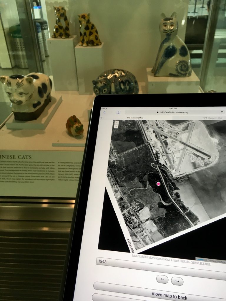 Photograph of a geolocated device, in the International Terminal Main Hall (c. 2018), facing the Caticons: The Cat in Art exhibition, with a 1943 aerial photo of the SFO campus.