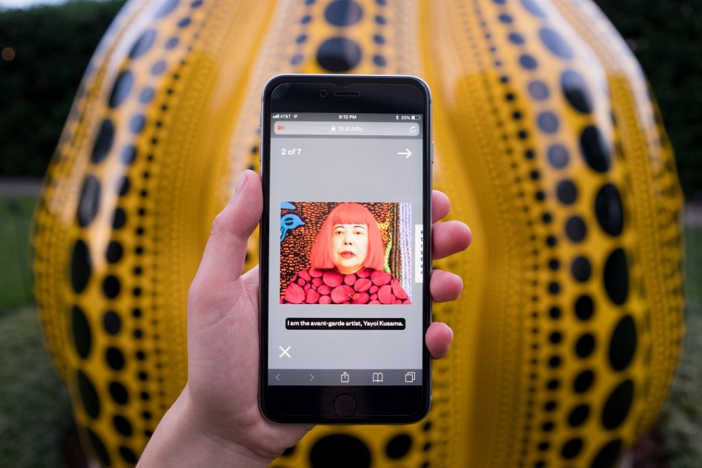 A mobile phone is held in front of a yellow pumpkin sculpture, playing a video of a woman onscreen. 